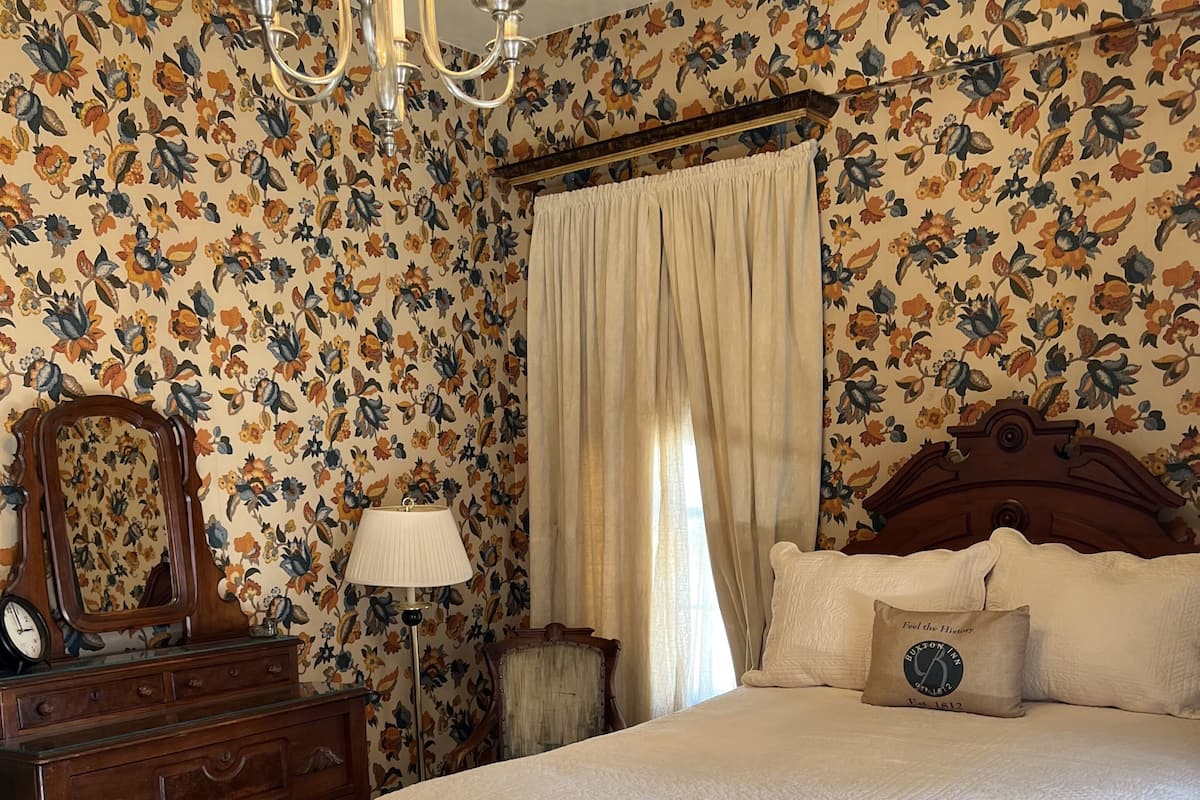 Une chambre à coucher avec un papier peint à motifs floraux, un lit en bois, une table de chevet avec une horloge et une commode.