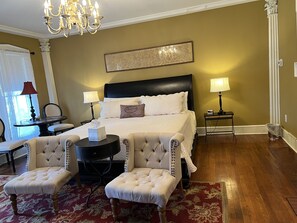 Una camera d'hotel con un letto grande, due comodini con lampade, un lampadario, un tappeto a motivi rossi e bianchi e due poltrone imbottite.