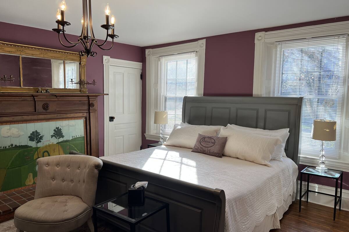 Une chambre à coucher comprenant un lit, une chaise, une table de chevet, une lampe, un miroir et un tableau accroché au mur.