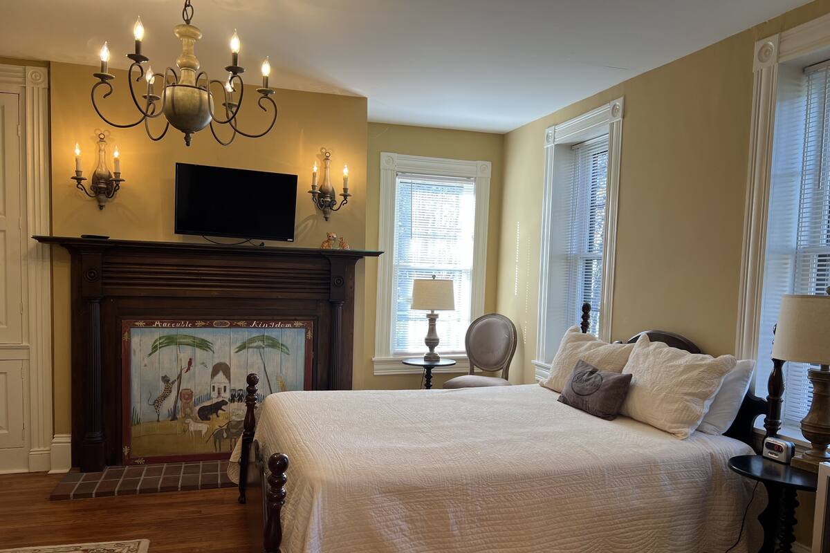Une chambre à coucher avec un grand lit, une cheminée ornée d’un tableau, un lustre et un téléviseur fixé au mur.