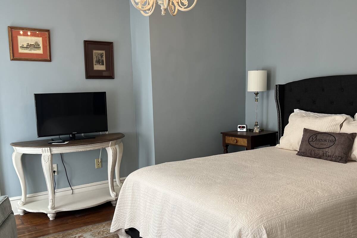 Une chambre à coucher avec un grand lit, un téléviseur posé sur une table blanche, un lustre, des tableaux encadrés au mur et une table de chevet avec une lampe.