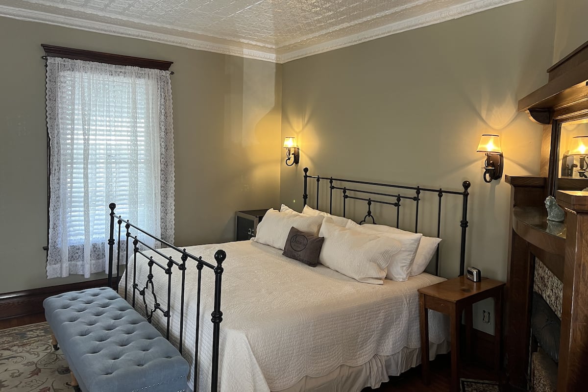 Une chambre à coucher comprenant un lit, un banc, deux tables de chevet, une commode, un miroir et une fenêtre avec des rideaux.