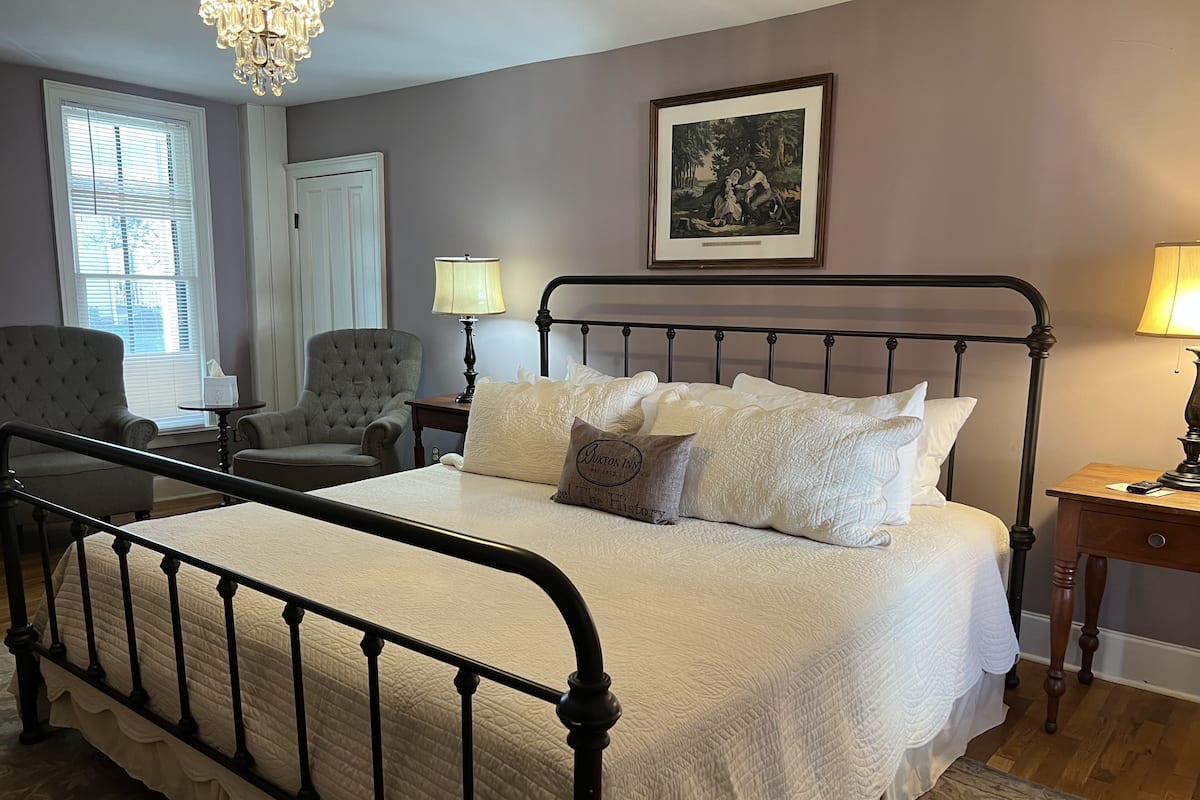 Une chambre à coucher bien rangée, avec un lit, deux chaises, un lustre, un tableau encadré et deux lampes de chevet.
