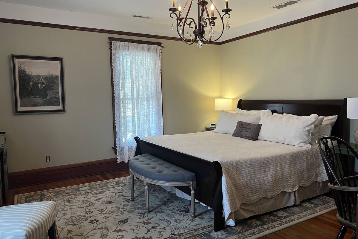 Une chambre à coucher avec un grand lit, une table de chevet, une chaise, un banc, un lustre et un tableau encadré au mur.