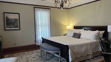 A bedroom with a large bed, a nightstand, a chair, a bench, a chandelier, and a framed picture on the wall.