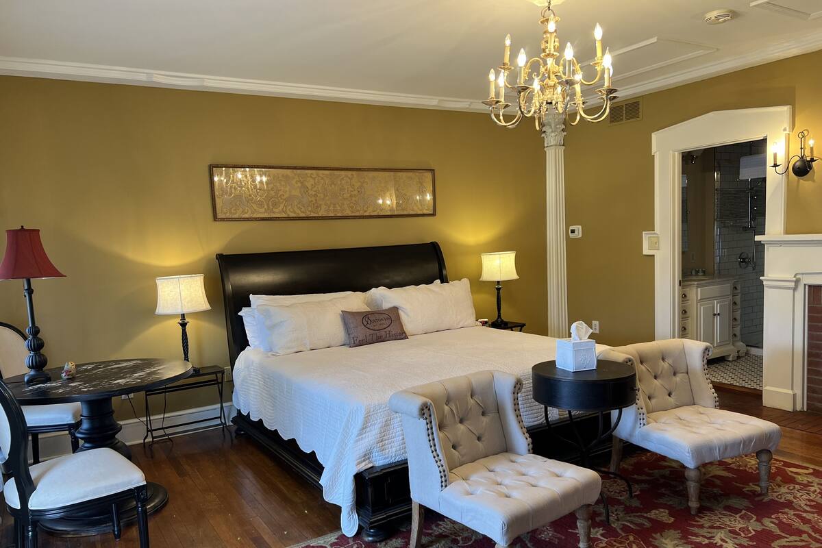 Une chambre élégamment aménagée, dotée d’un grand lit, de deux tables de chevet avec des lampes, d’un lustre et d’un coin salon avec des fauteuils et une petite table.