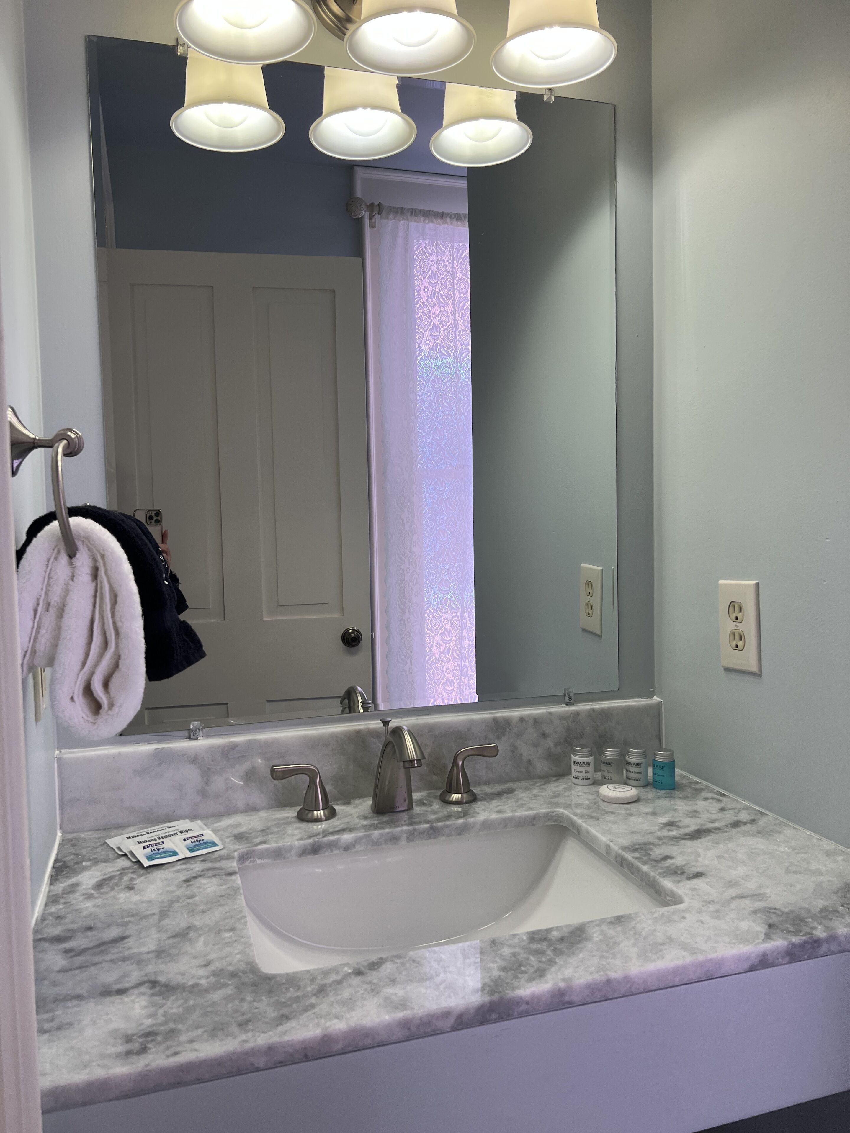 A bathroom with a white sink, three faucets, and a large mirror above it. There are towels hanging on a rack to the left, and a door with a white curtain in the background.