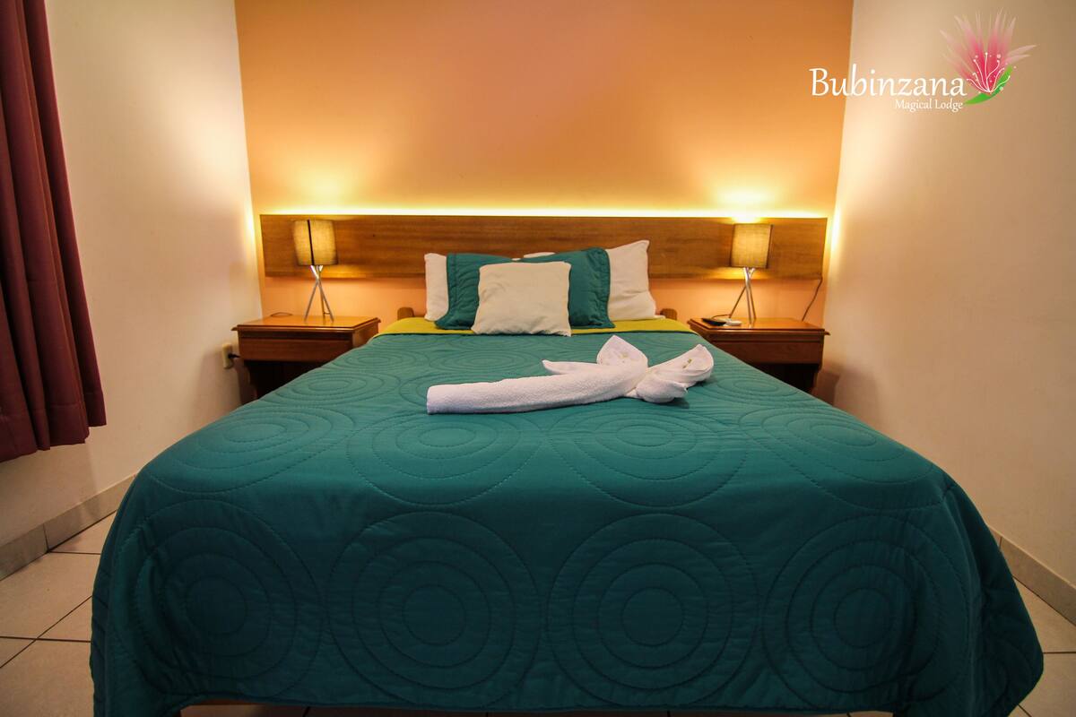 Ein ordentlich gemachtes Bett mit einem saphirblauen Bettüberwurf und weißen Kissen, zwei Nachttischlampen und ein sauber gefaltetes Handtuch auf dem Bett.