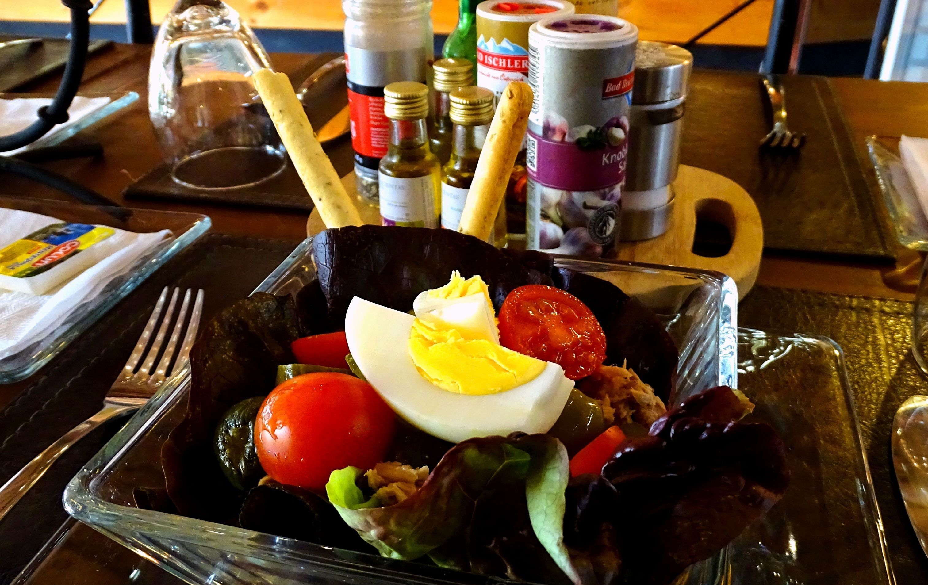 A salad with a boiled egg, tomato, and lettuce on a table with condiments and a fork.