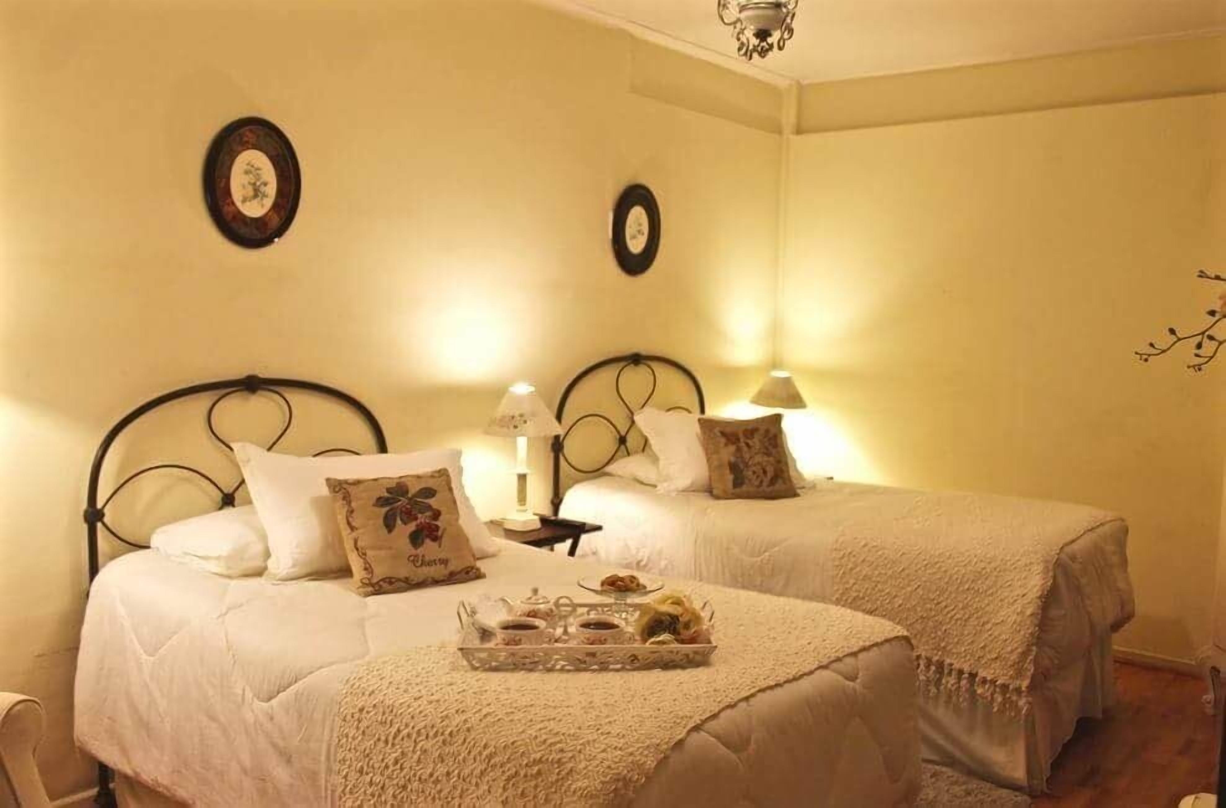 A room with two beds, each with a headboard, a bedside table with a lamp, and a tray with food items.
