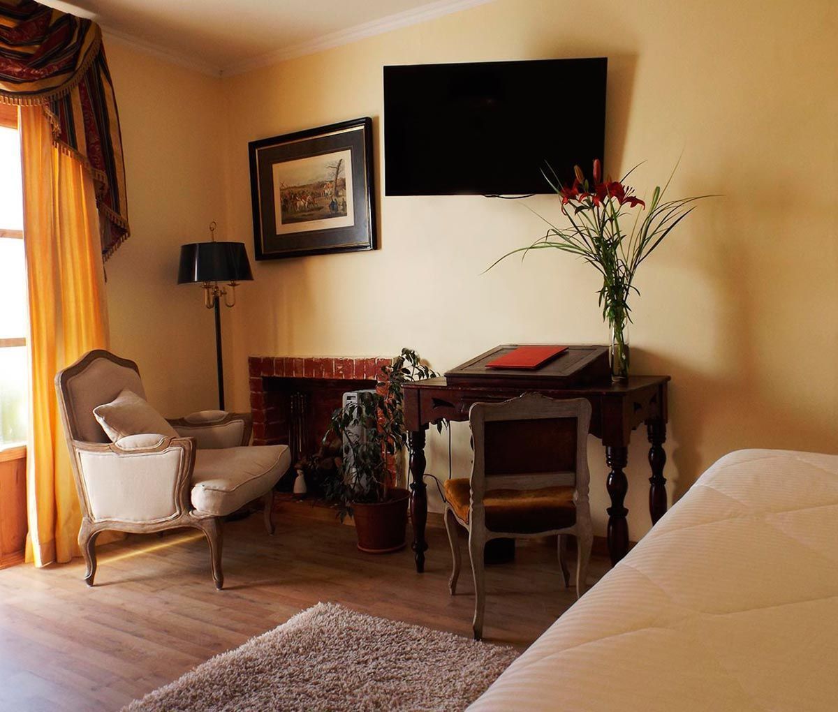 Ein Hotelzimmer mit einem Bett, zwei Sesseln, einem Schreibtisch mit Blumen in einer Vase sowie einem flachbildfernseher, der an der Wand montiert ist, und einem gerahmten Bild über dem Fernseher.