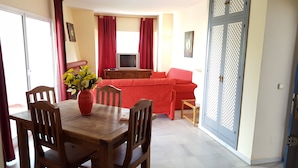 A dining area with a wooden table and chairs, a vase of flowers, and a red sofa in the background. - Resort Medina del Zoco (Mijas)
