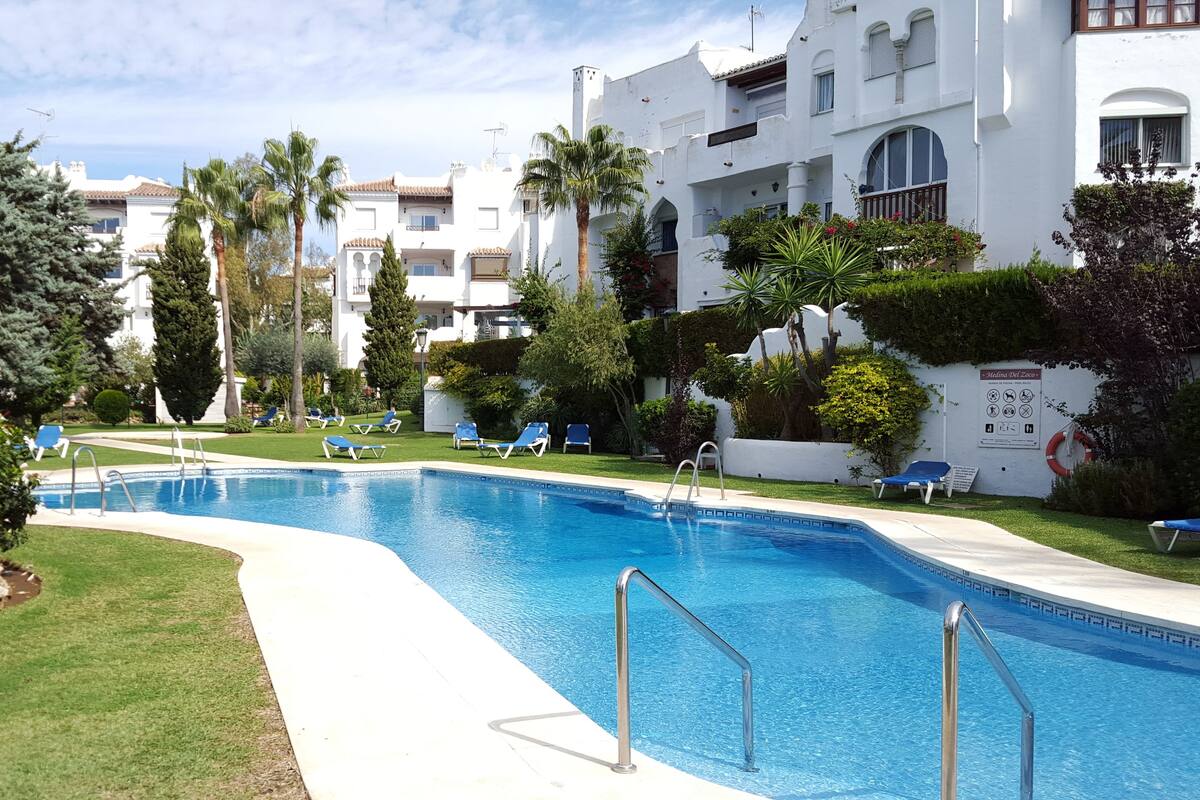 A swimming pool with lounge chairs, surrounded by greenery and white buildings with balconies.