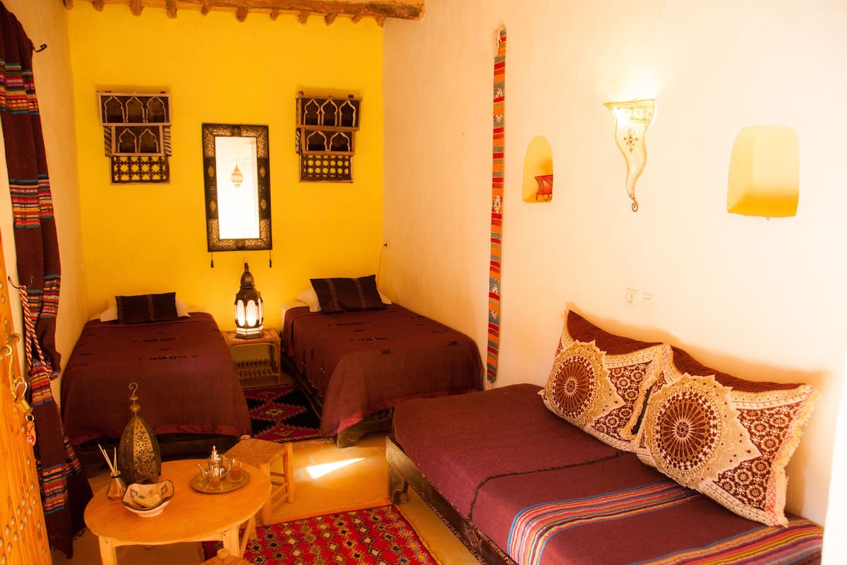 A room with two beds, a small table with a teapot, and a wall-mounted lamp.