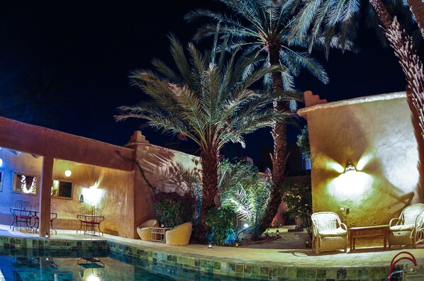 An outdoor pool area with chairs and tables, surrounded by palm trees and a building with warm lighting.