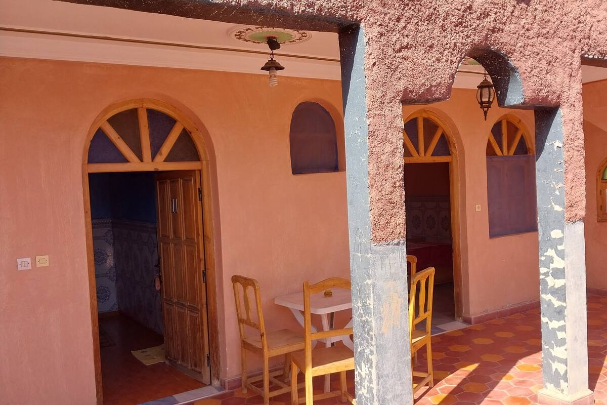Un espace extérieur aménagé, abrité et doté de chaises en bois et d’une table, relié à un bâtiment par des portes en arc de cercle et dont le sol est recouvert de carreaux.