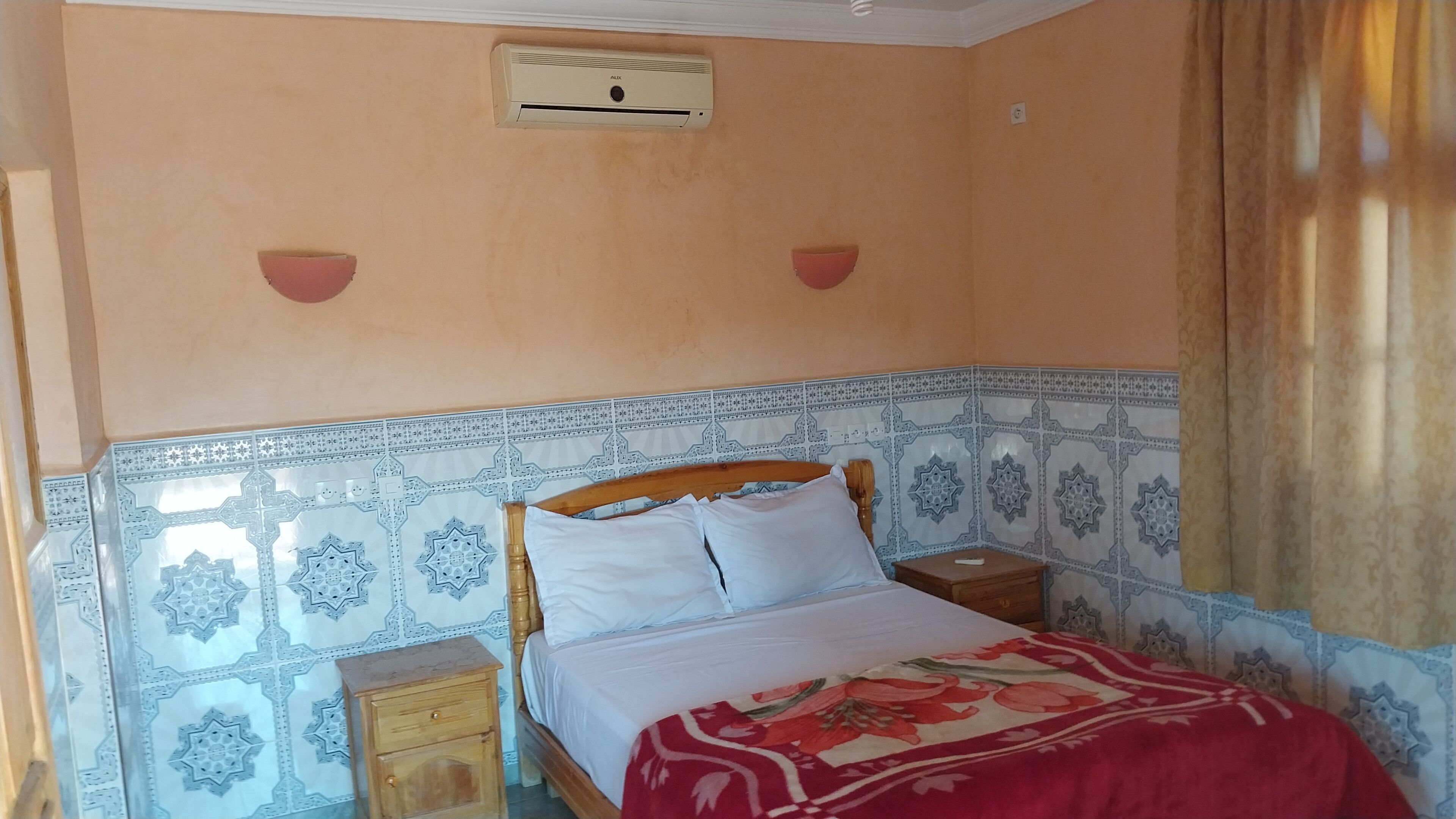 Een slaapkamer met een bed, nachtkastjes, een wandairconditioner en tegels met een patroon.