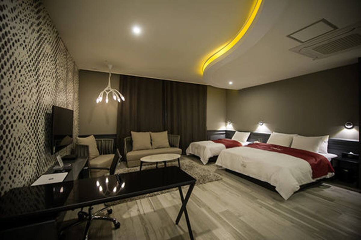 A modern hotel room with a sofa, two beds, a desk, and a TV.