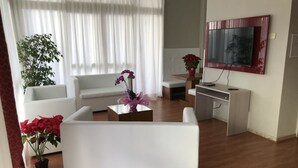 Ett modernt vardagsrum med en vit soffa, ett soffbord med blommor, en tv som är monterad på väggen och ett stort fönster med tunna, genomskinliga gardiner.