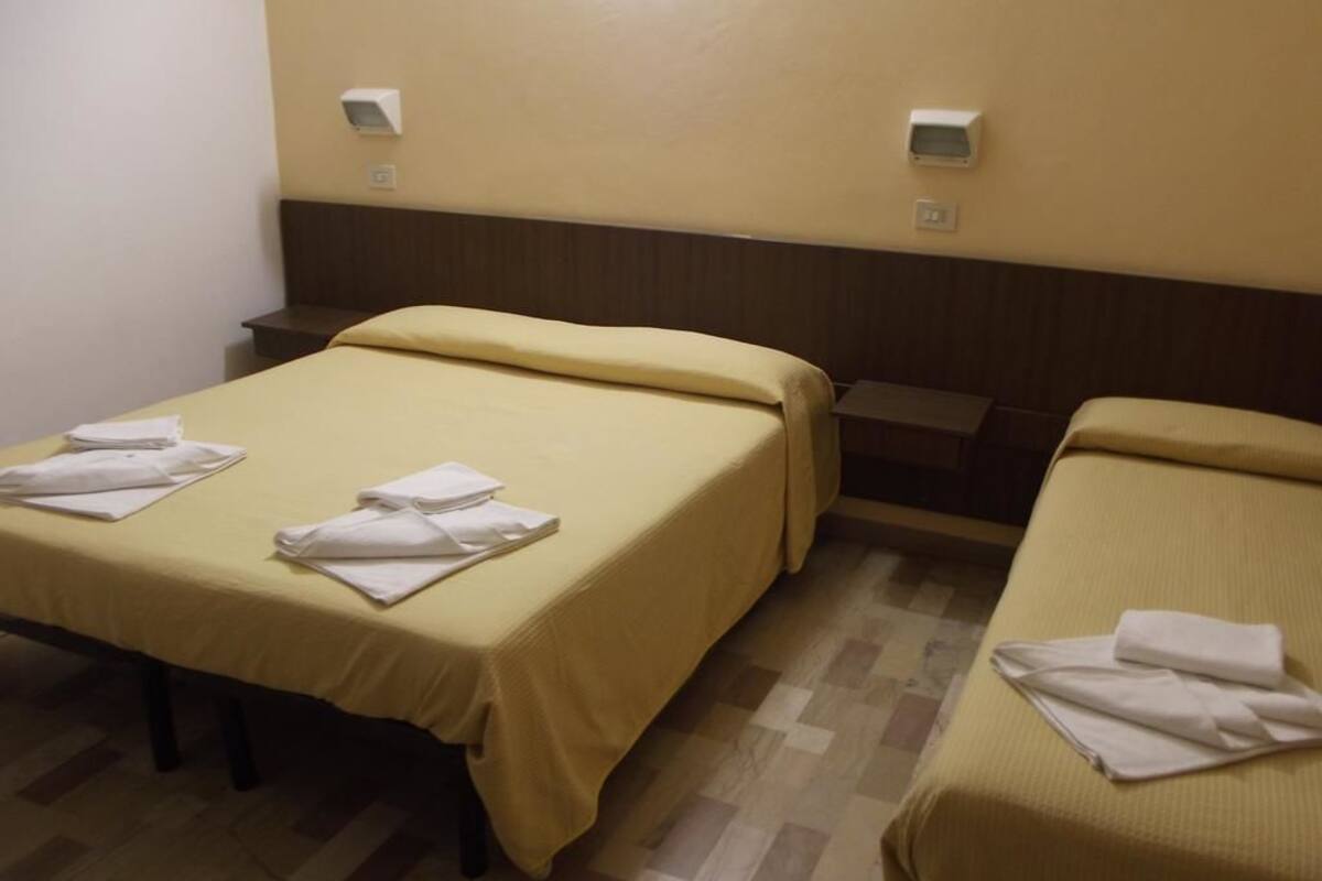 A hotel room with two beds, each with a yellow bedspread and white towels. There are wall-mounted light switches and a small table between the beds.