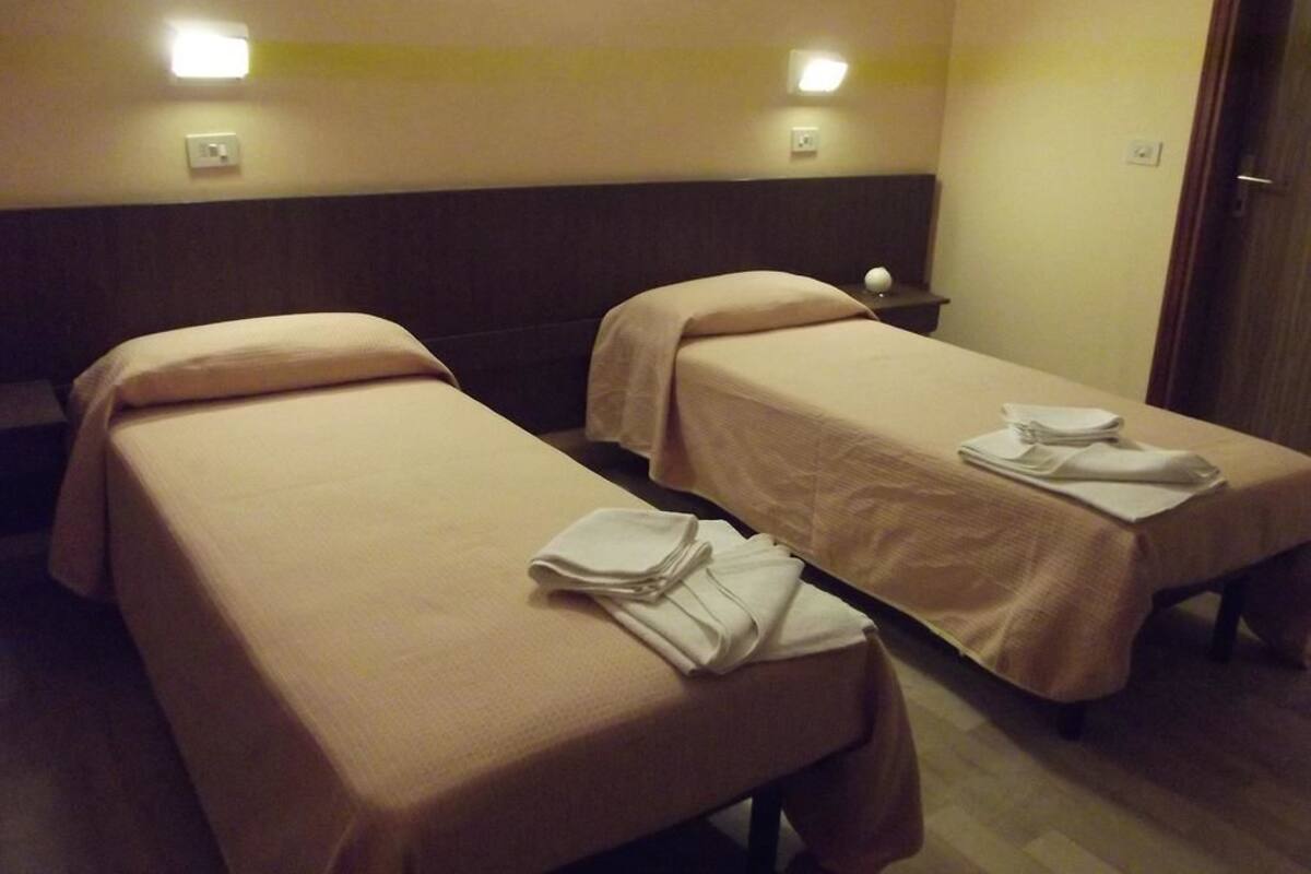 Two single beds with beige linens and folded towels, a bedside table, and a wall-mounted light fixture.