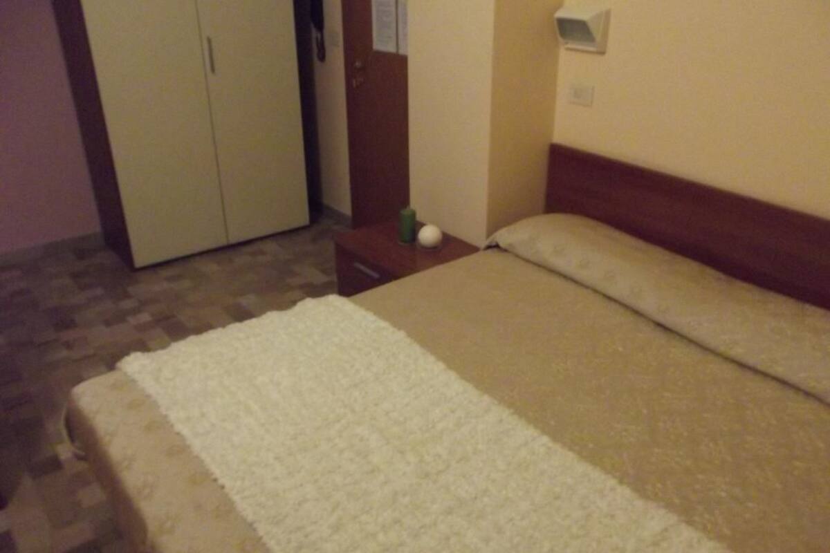 A bedroom with a bed, a nightstand, a wardrobe, and a wall-mounted shelf.