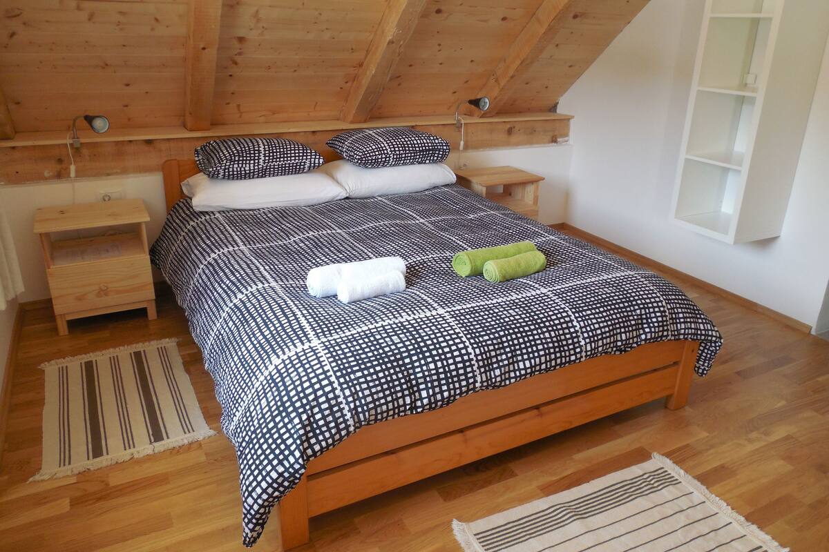 Una cama de madera con colcha a cuadros, dos almohadas, dos mesitas de noche y una ventana con repisa.