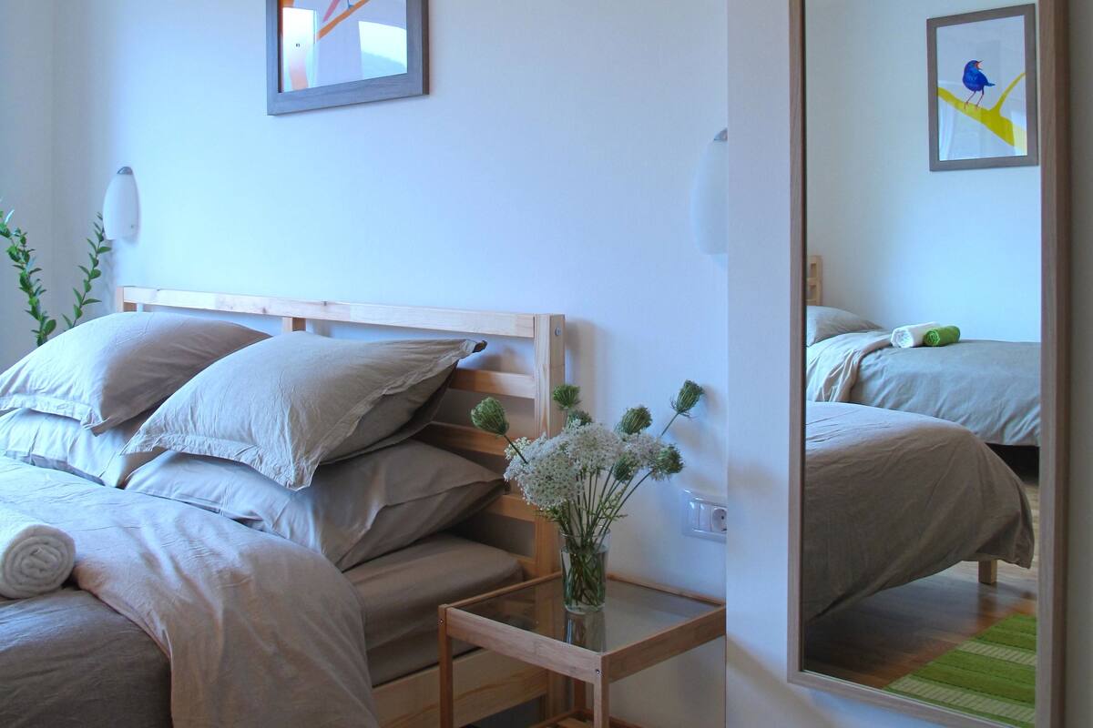 Un dormitorio con cama, almohadas, una mesita de noche con un florero, un espejo y dos cuadros en la pared.