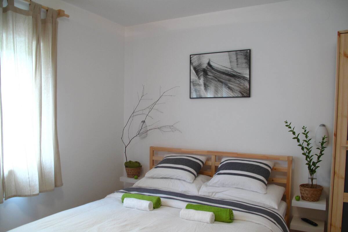 Una cama bien hecha con ropa de cama a rayas, un cuadro abstracto colgado en la pared, una planta en maceta y una mesita de noche con un control remoto.