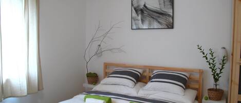 A neatly made bed with striped bedding, a wall-mounted abstract painting, a potted plant, and a bedside table with a remote control.