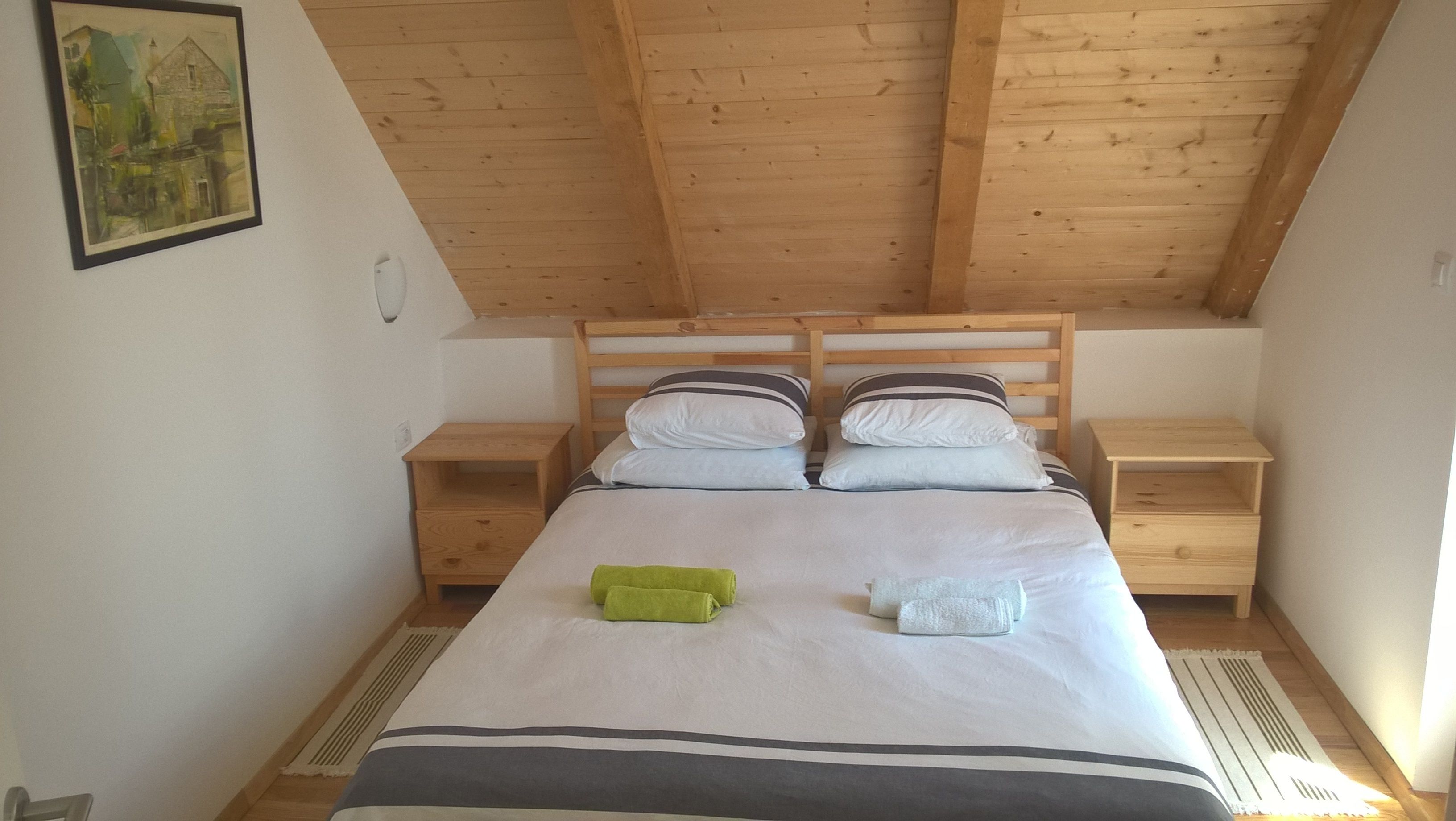 A bedroom with a wooden ceiling, a bed with white bedding, two bedside tables, and a painting on the wall.