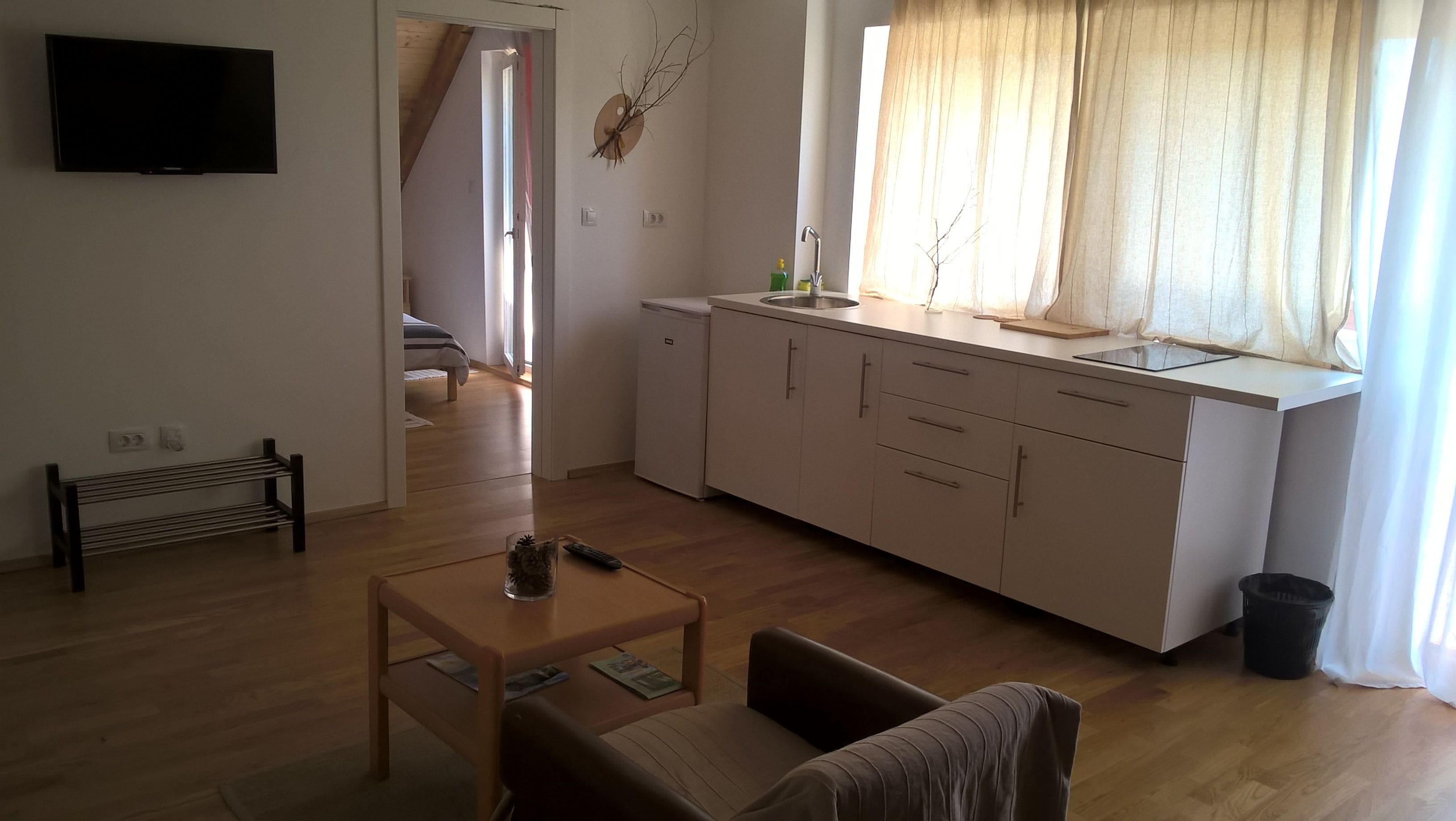 A living room with a sofa, a table, and a TV. There is a kitchen area with a sink and cabinets. A window with curtains is visible.
