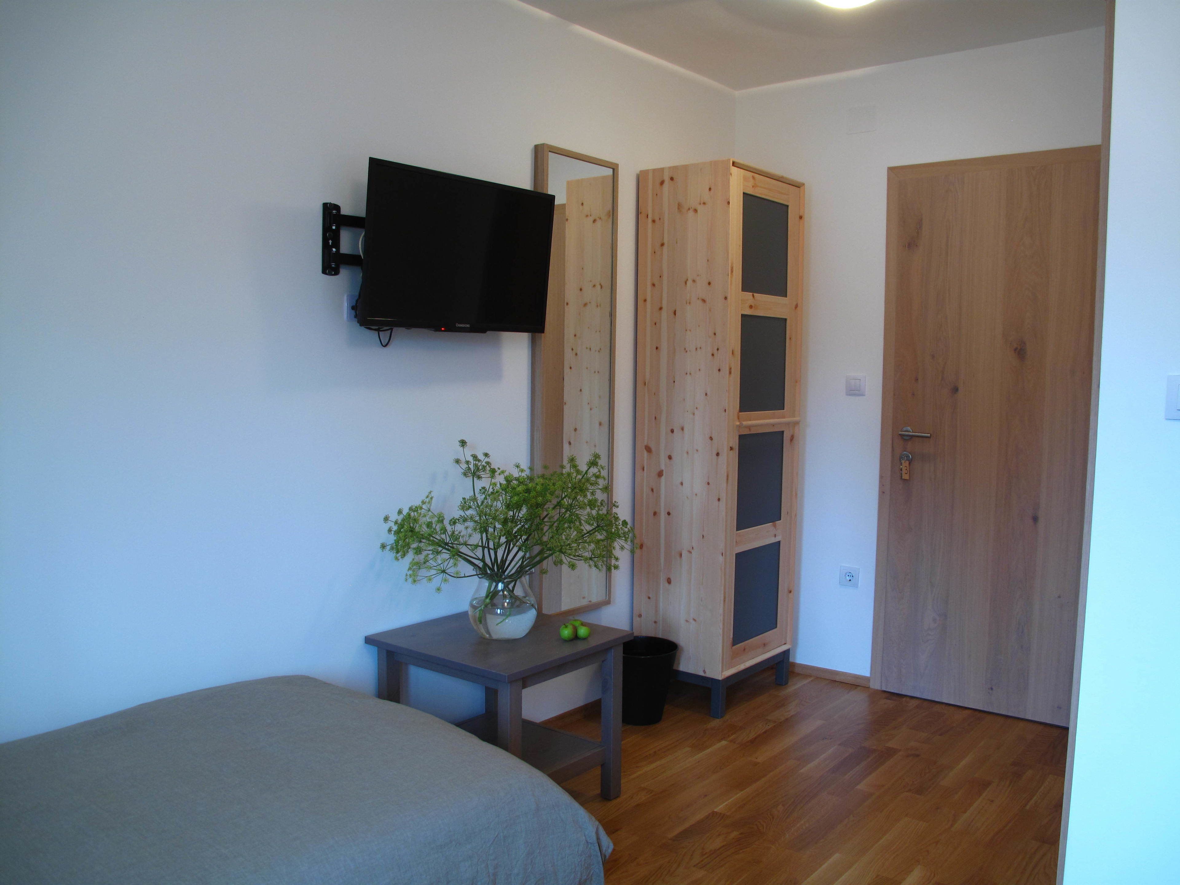 A bedroom with a bed, a TV mounted on the wall, a wooden wardrobe, and a small table with a plant.