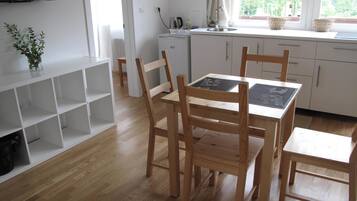 A modern kitchen with white cabinets, a wooden dining table, and chairs. There is a TV mounted on the wall, a window with curtains, and a plant on the left.