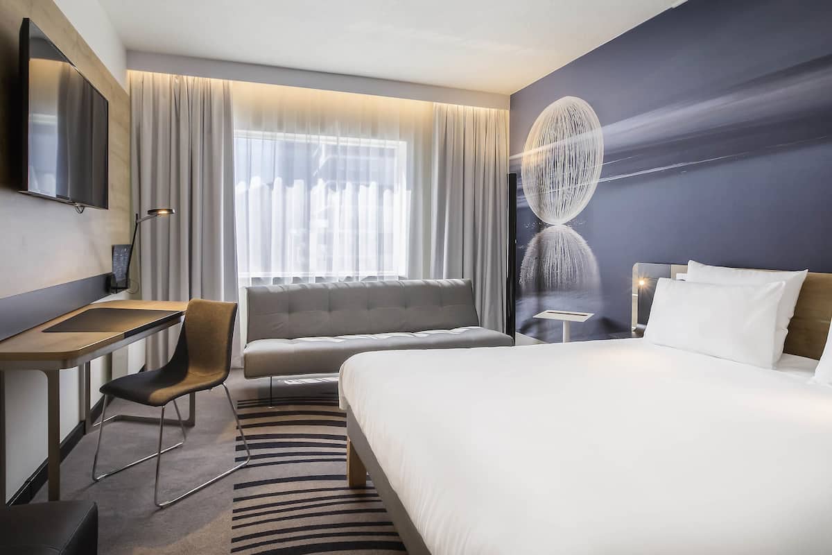A hotel room with a bed, a desk, a chair, a TV, and a large window with curtains.