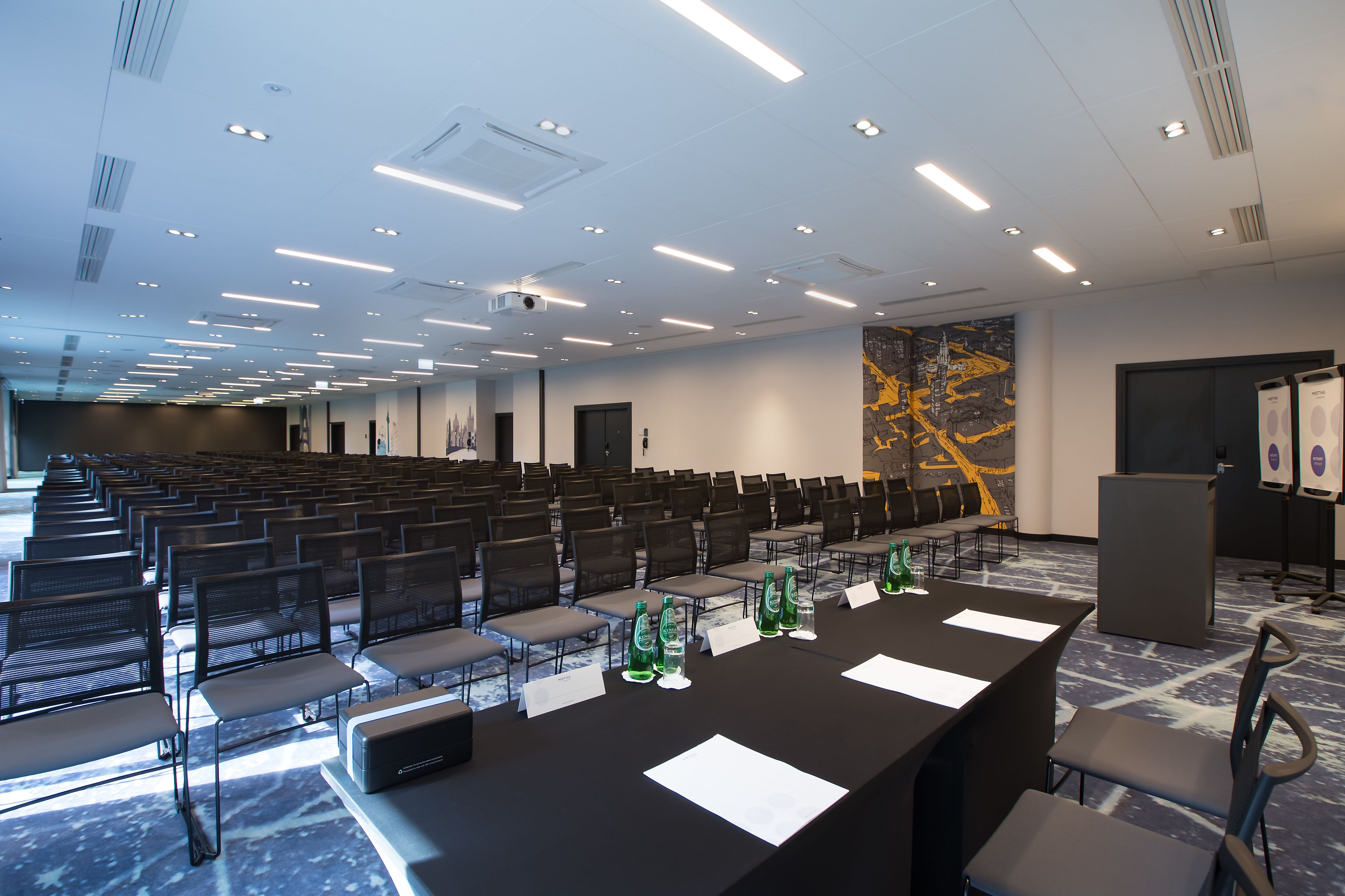 A conference room with rows of chairs, a long table with nameplates, and a projector screen.