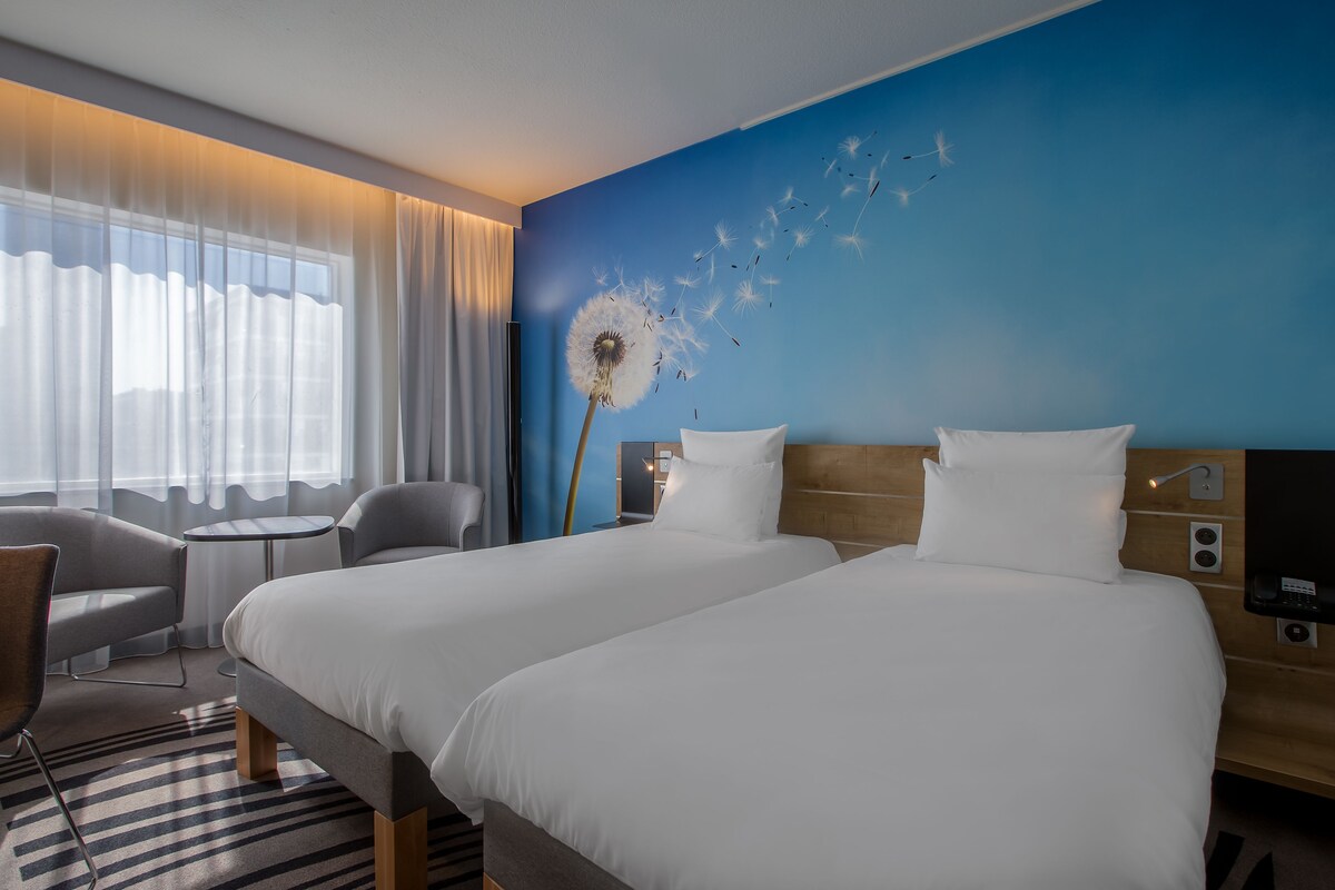 Habitación de hotel con dos camas, un escritorio, una silla y un ventanal con cortinas.