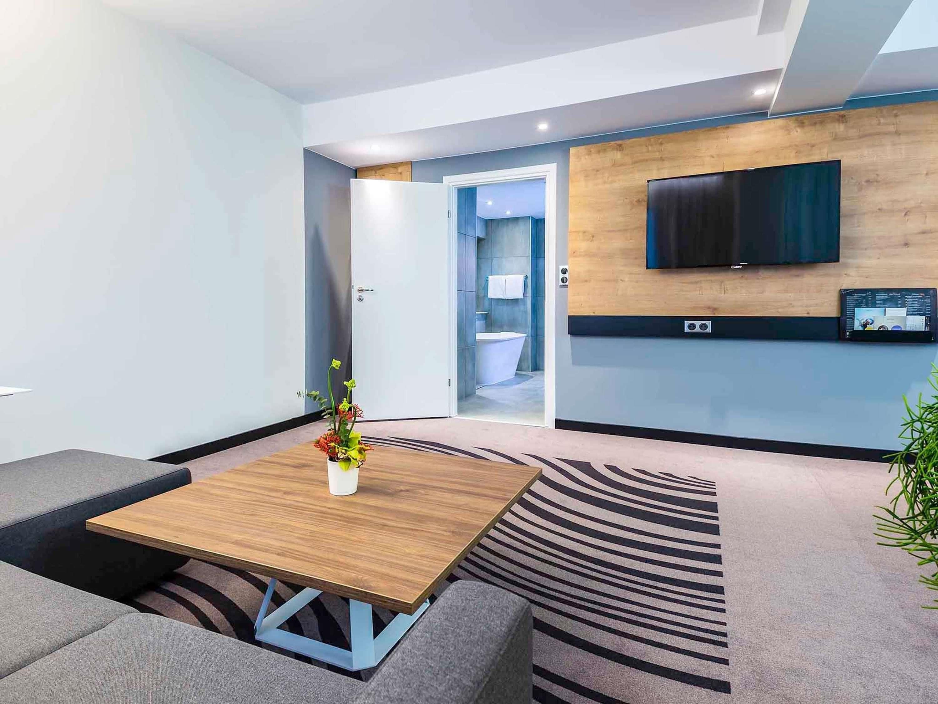 A modern hotel room with a flat-screen TV mounted on the wall, a wooden coffee table, grey sofas, and a patterned carpet.