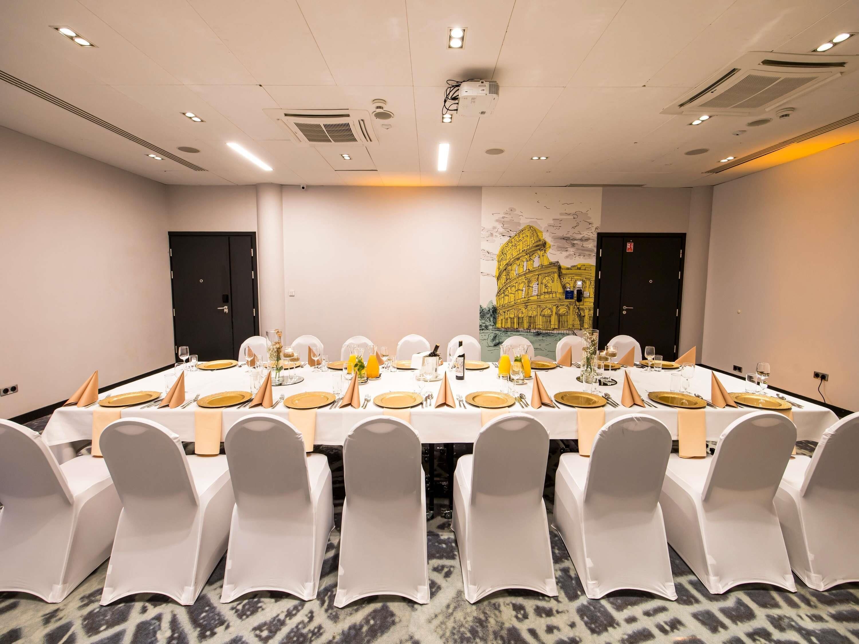 Un salón de conferencias con una larga mesa dispuesta para una reunión, sillas blancas y un gran mural del Coliseo.