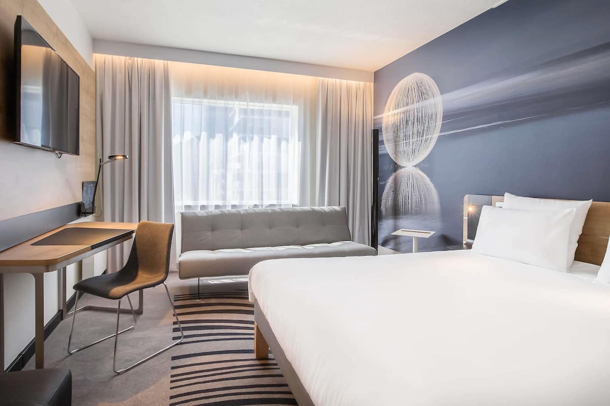 Quarto de hotel moderno com cama, sofá, escrivaninha, cadeira e televisão.