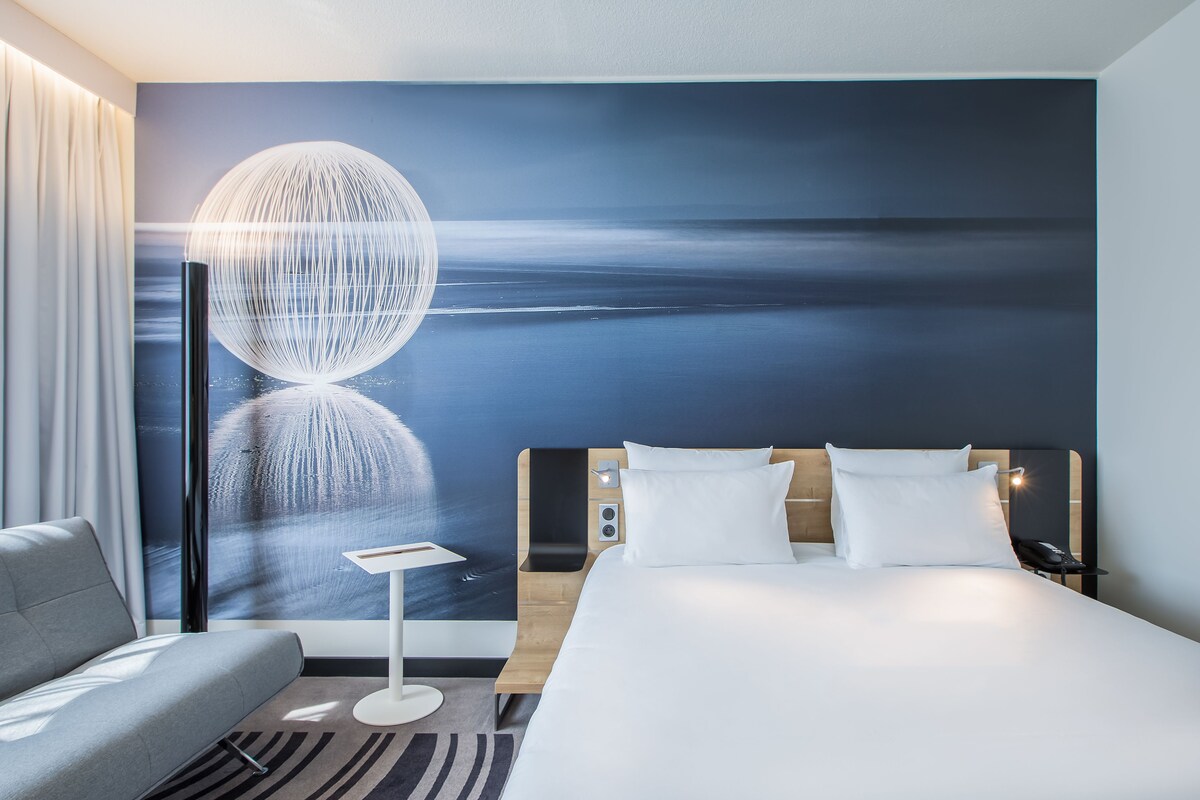 Una habitación de hotel moderna con una cama grande, un sofá, una mesita de noche y un mural en la pared con un cielo nocturno.