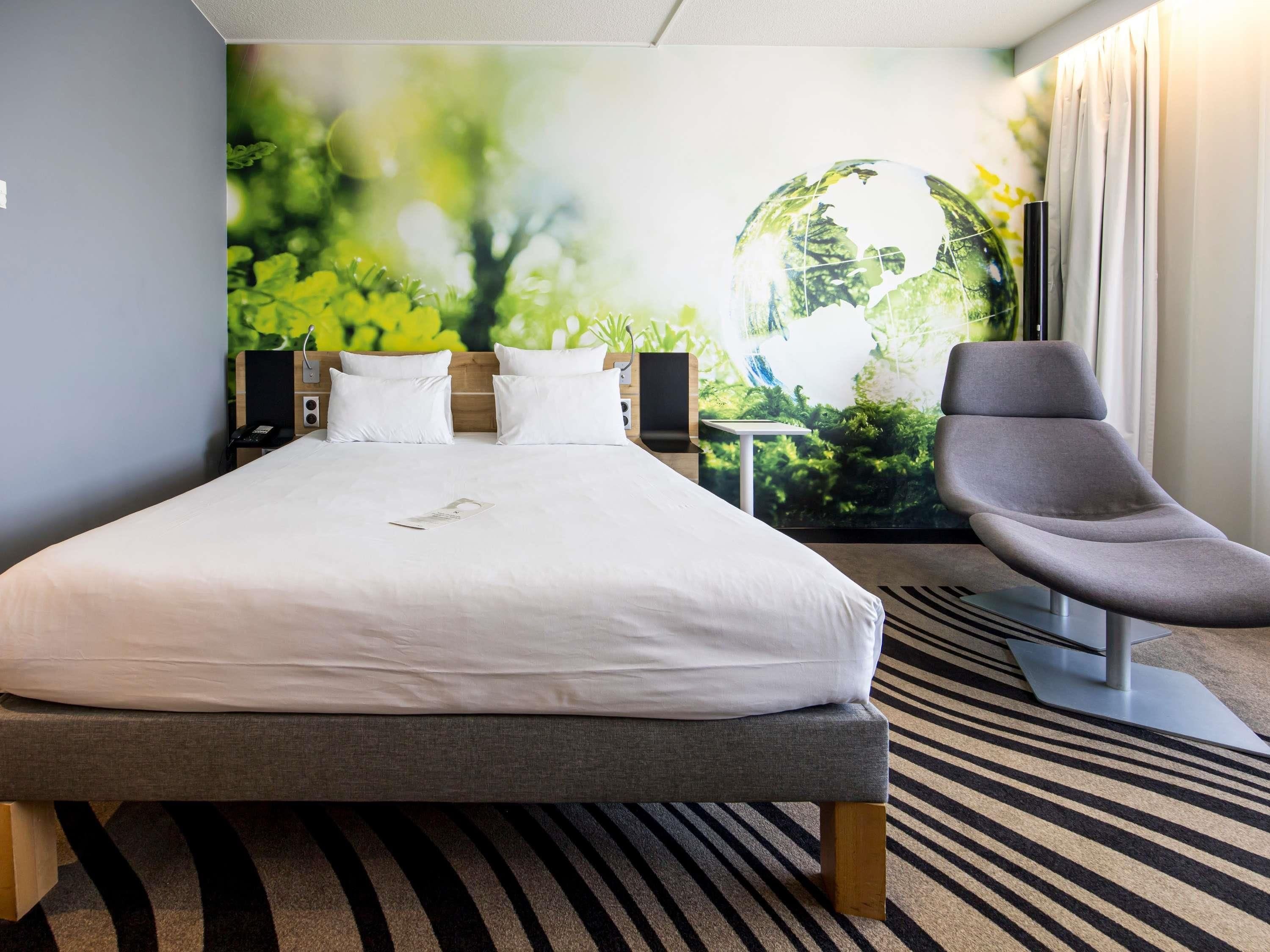 A hotel room with a large bed, a grey armchair, a wall mural, and a small table.