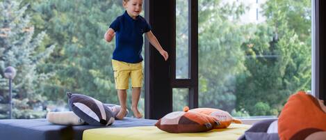 Un niño de pie sobre una cama con almohadas, una ventana y follaje visible desde el exterior.