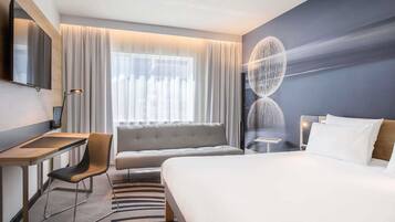 A hotel room with a bed, a desk, a chair, a sofa, a TV, and a large window with curtains.