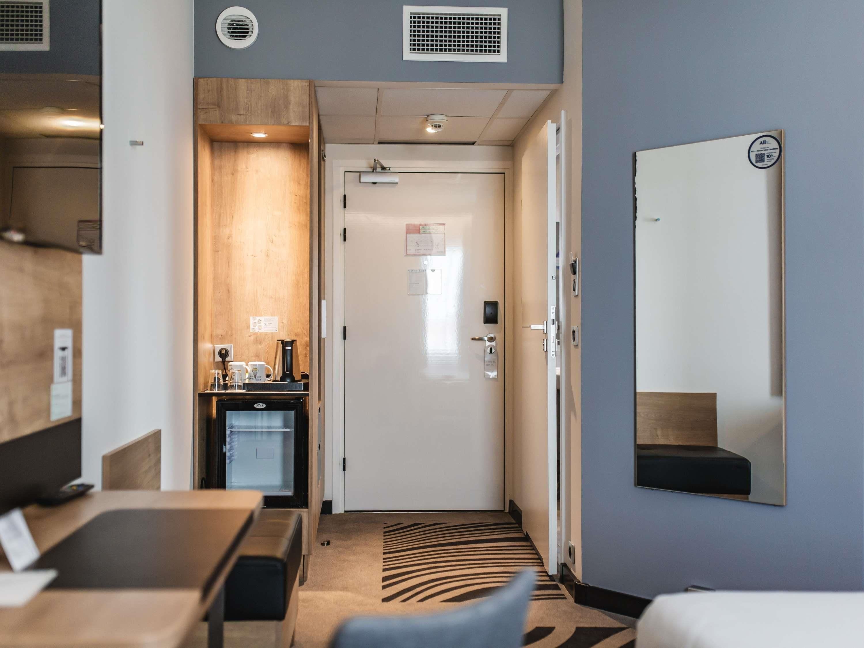 A hotel room with a desk, a minibar, a mirror, and a door leading to another room.