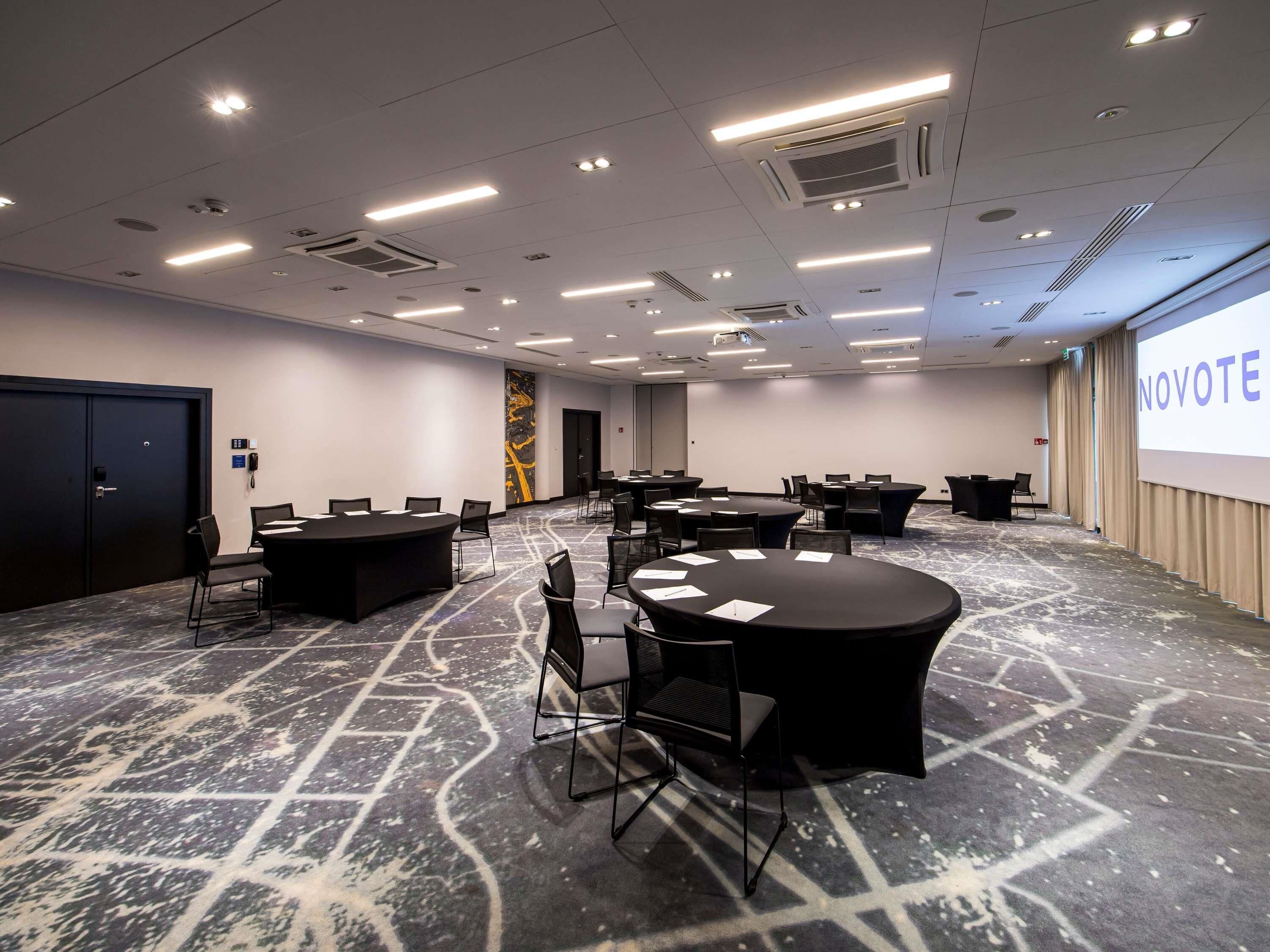Sala de conferências com uma tela grande, várias mesas e cadeiras.