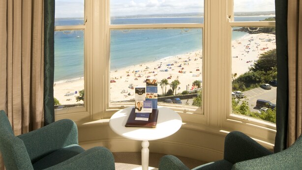 Una habitación con vista a la playa, que incluye dos sillones tapizados en verde, una mesita blanca con un libro y un menú, y una ventana con cortinas color beige.