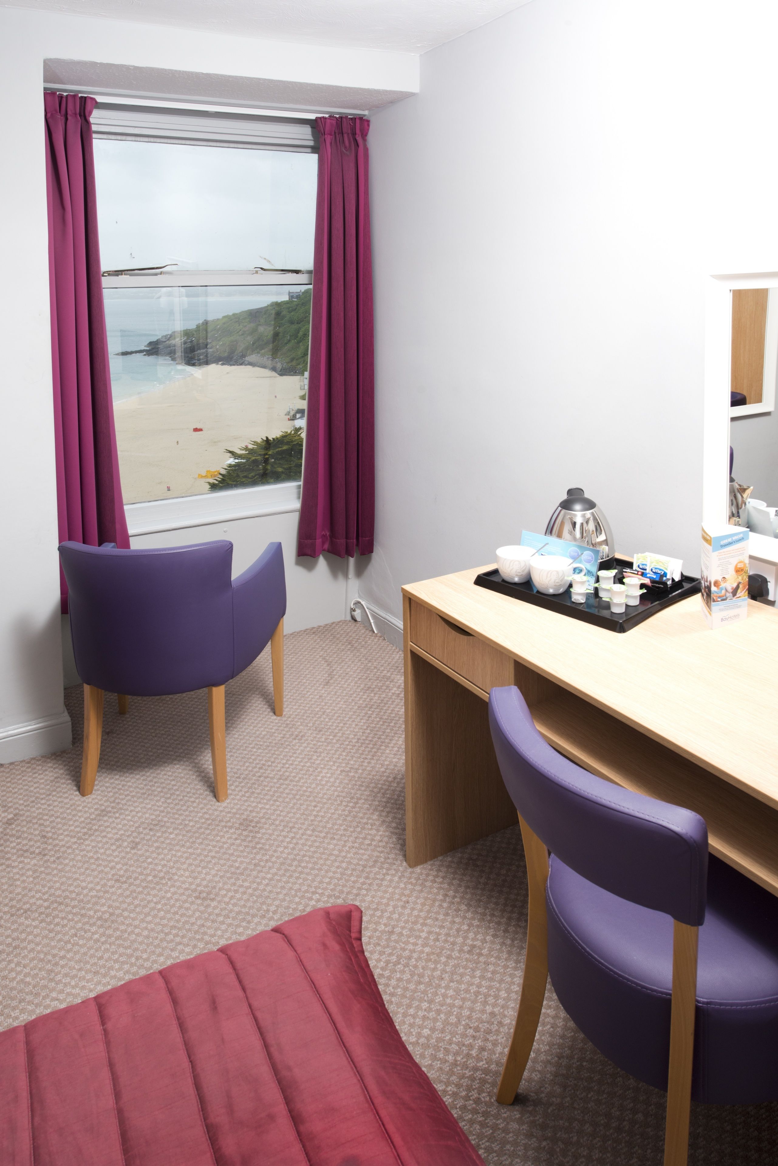 A room with a wooden desk, purple chairs, a window with curtains, and a view of the beach.
