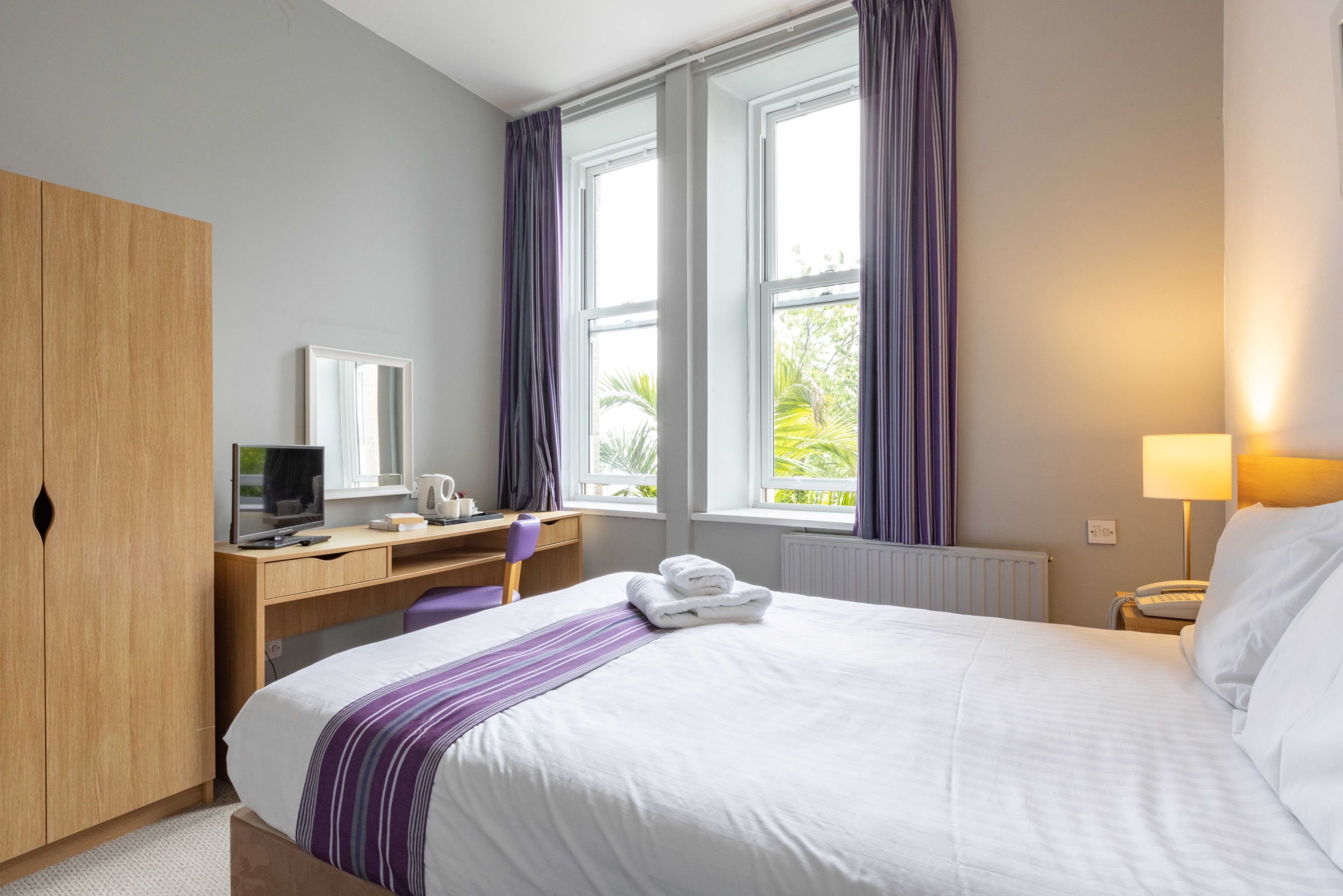 A hotel room with a bed, a desk, a chair, a wardrobe, and a window with curtains.
