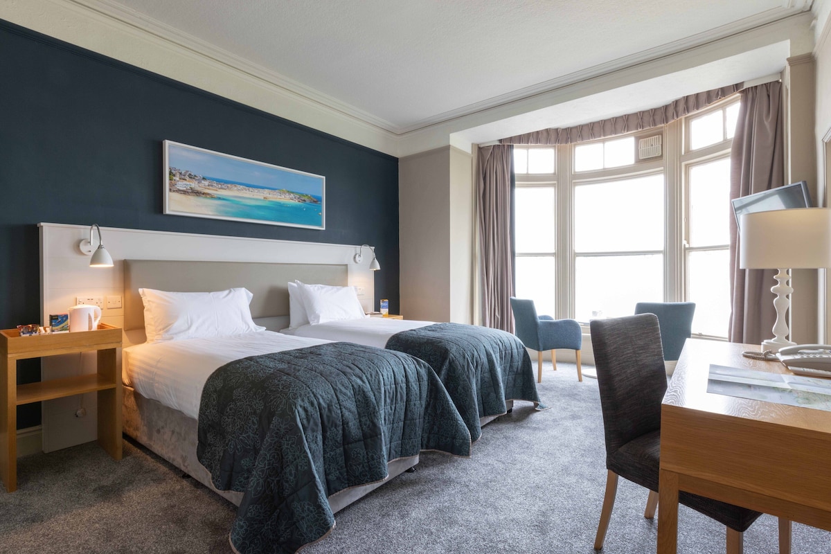 A hotel room with two beds, a desk, a chair, and a painting on the wall.
